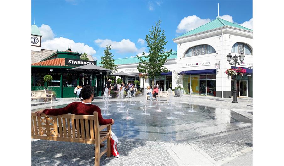 Outdoor seating area at McArthurGlen West Midlands Designer Outlet, featuring a central fountain, benches, shoppers, and storefronts including Starbuc