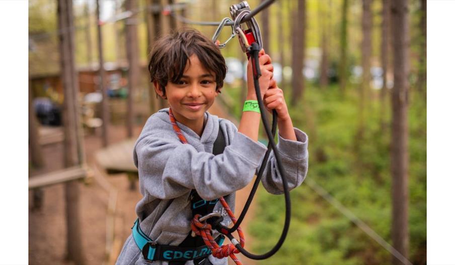 A child wearing a safety harness and gripping a zip line handle while moving through an outdoor high ropes course set in a forested area.