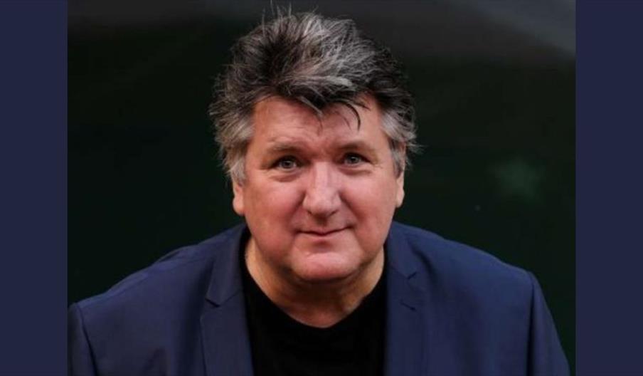 Bob Mills wearing a dark navy blazer over a black shirt is positioned against a dark background.