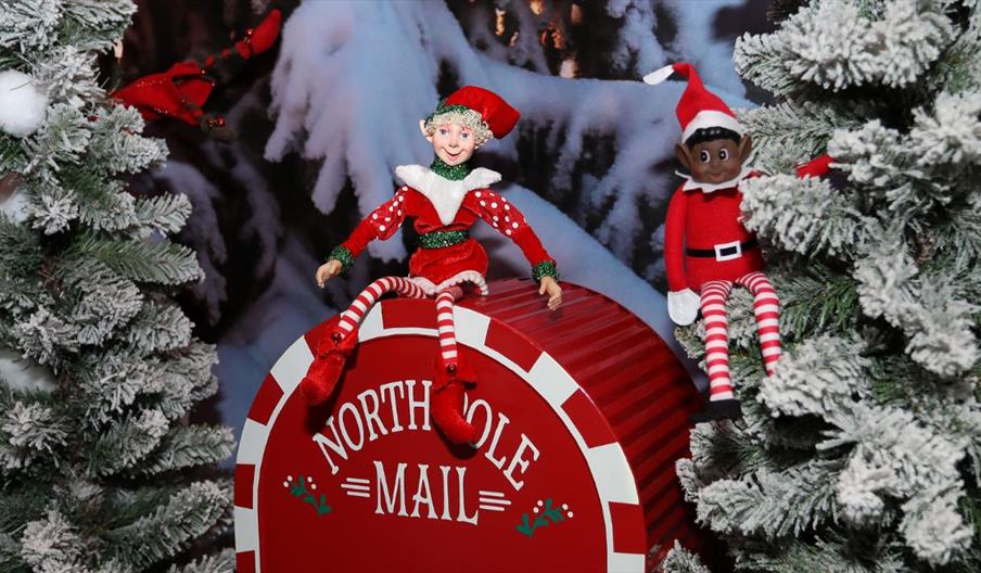 Image 1: Festive scene with two Christmas elves sitting on a red North Pole Mail barrel surrounded by snow-covered trees.
Image 2: Father Christmas se