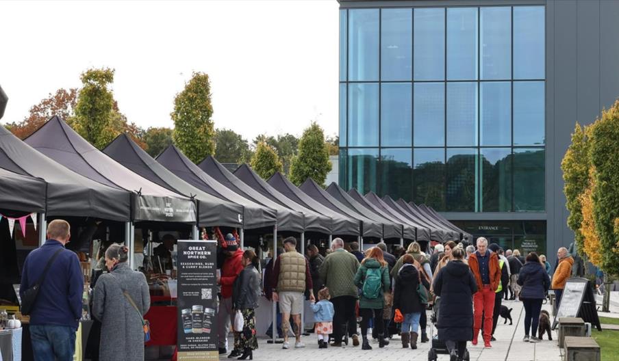 Outdoor artisan market at World of Wedgwood, with rows of black‑topped stalls and visitors walking between them. A modern glass-fronted building stand