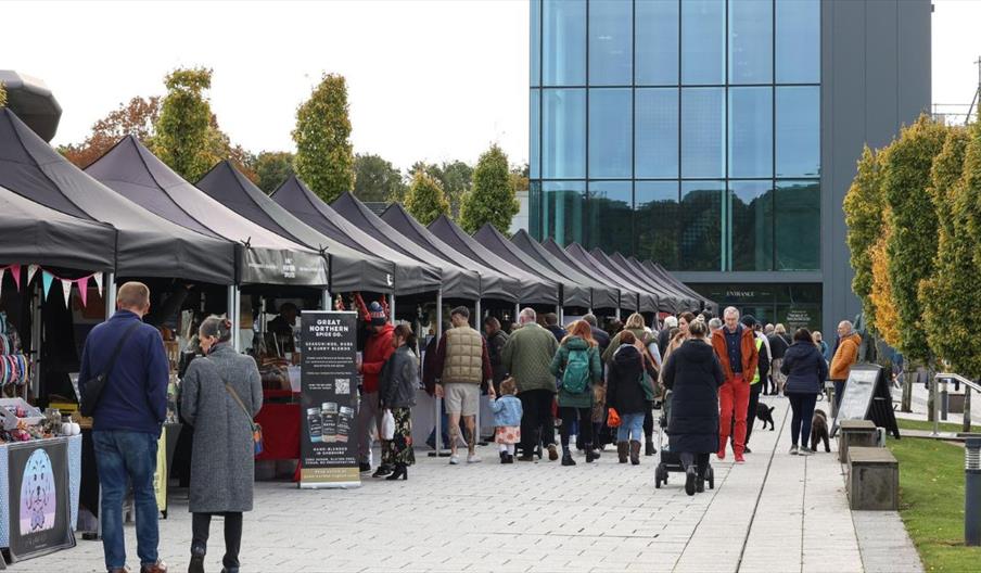 Outdoor market with multiple black canopy stalls lined up along a paved walkway. People are walking and browsing the stalls, which display various goo