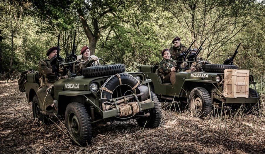 Two vintage military jeeps parked in a woodland setting with people in uniform seated inside.