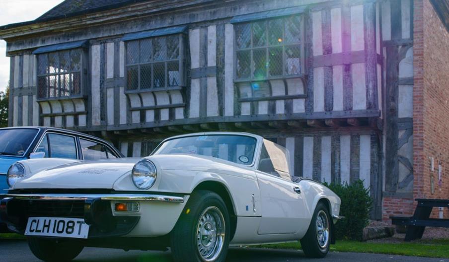 A white classic convertible car parked in front of a historic timber‑framed building, with another vintage car beside it.