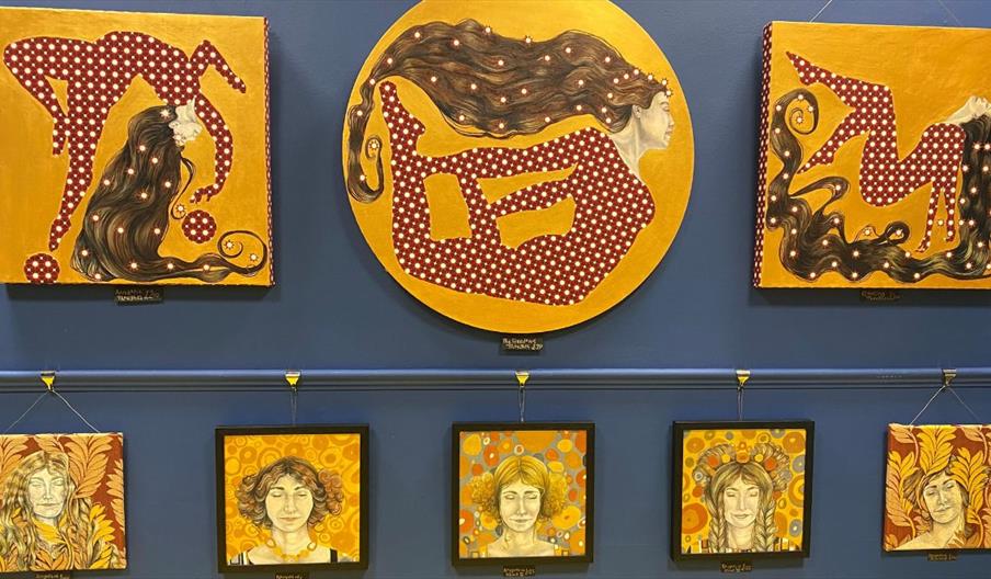 A display of vibrant, patterned artworks on a blue wall, featuring stylized figures with flowing hair in red and gold tones, alongside smaller framed