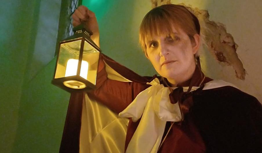 A person dressed in a historical or theatrical costume holds a lantern with a glowing candle inside, casting warm light. The outfit includes a dark cl