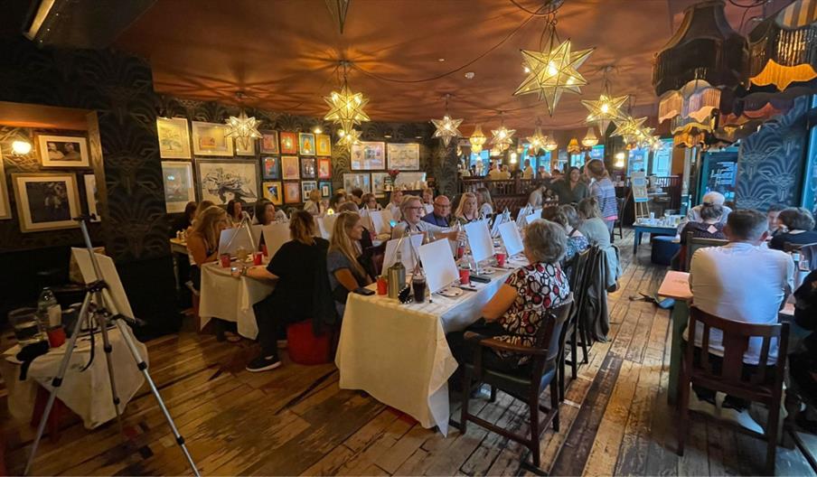 People taking part in a guided painting event, seated at long tables with canvases and art materials in a warmly lit venue decorated with framed artwo