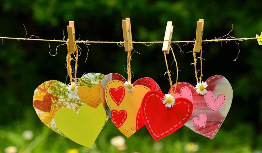 Four colourful paper heart decorations hanging from a string with clothespins, set against a blurred green garden background.