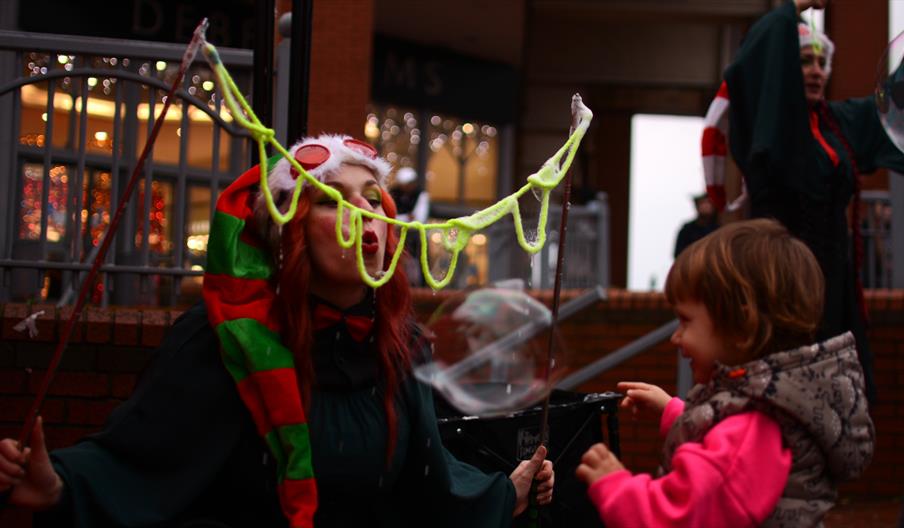 A woman in Christmas scarf and hat blows a huge bubble as a child looks on