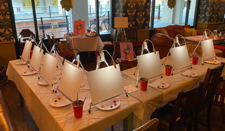 A cosy venue set up for a guided painting event, with tables arranged with blank canvases on easels, paint palettes, brushes, and drinks, ready for pa