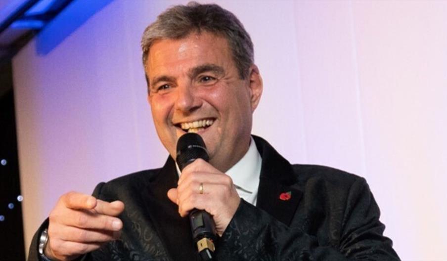 Dave Bryon wearing a dark patterned jacket and holding a microphone while speaking on stage, with a light-colored backdrop and some decorative lights