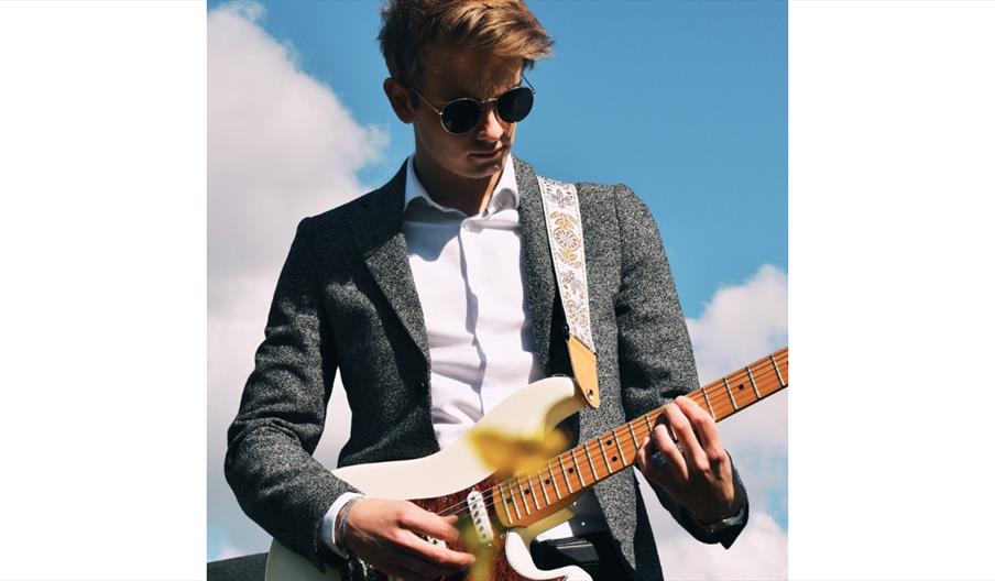 A person playing an electric guitar outdoors against a bright blue sky with white clouds.