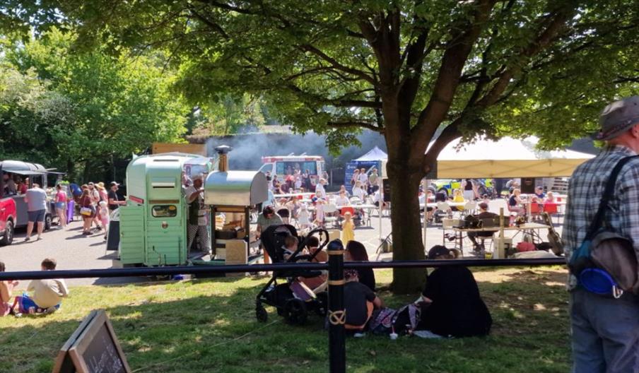 A busy outdoor event at a castle setting on a sunny day. Families are sitting and relaxing on grass under leafy trees while food stalls and trailers o