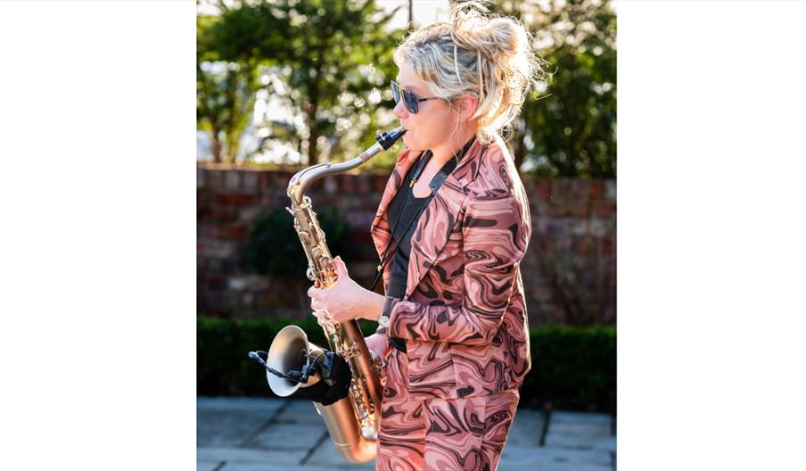 A musician playing a saxophone outdoors in daylight, wearing patterned clothing and sunglasses.