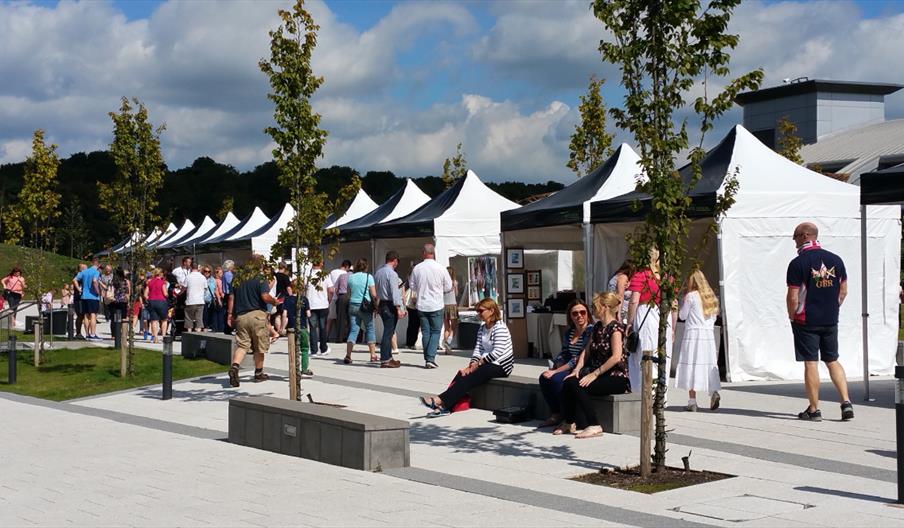 An outdoor artisan market set along a walkway with a row of white marquee stalls on a sunny day. Visitors are browsing stalls displaying crafts and ar