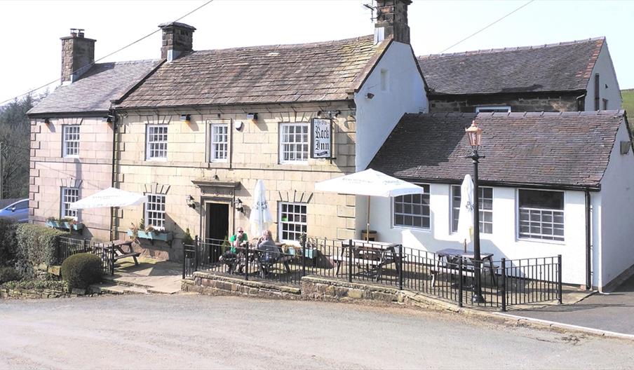 Image shows the exterior of Ye Olde Rock Inn, on a sunny day, with a few customers sitting at the tables outside