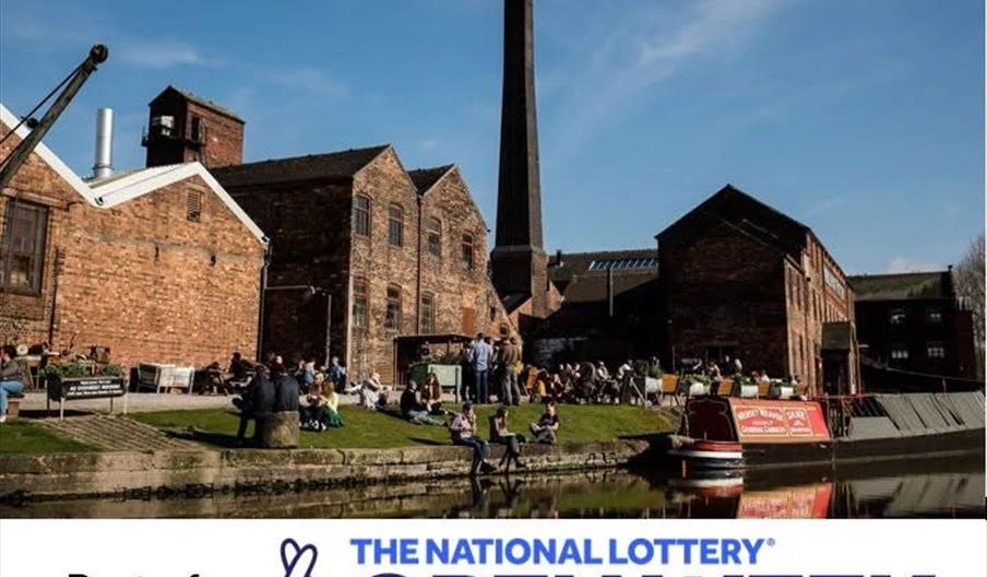 An image of Middleport Pottery on a glorious day, with visitors sitting on a patch of grass beside the canal, a narrowboat moored up, and the historic