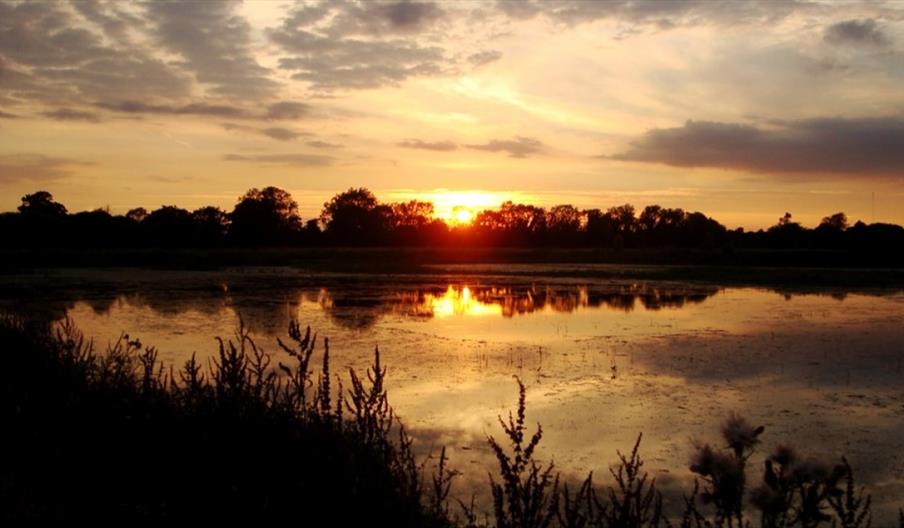 Image shows a sunset at Middleton Lakes, Staffordshire, with the sun dipping behind the trees and casting a pretty reflection on the lake