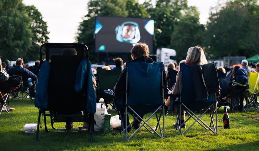 Film fans sit in garden chairs and watch a film on a big screen in a park