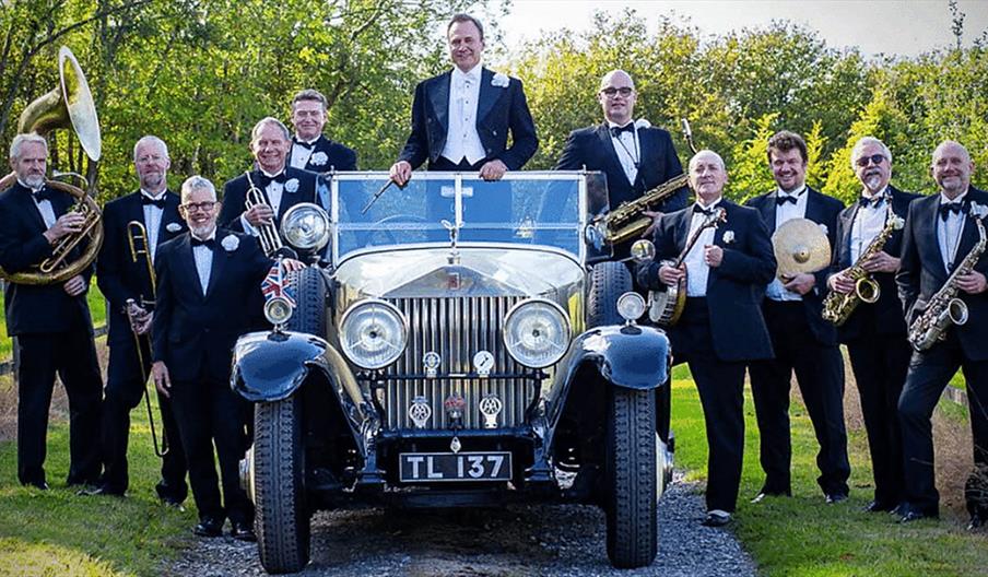 The musicians of the Pasadena Roof Orchestra hold their instruments, as one stands in an old car