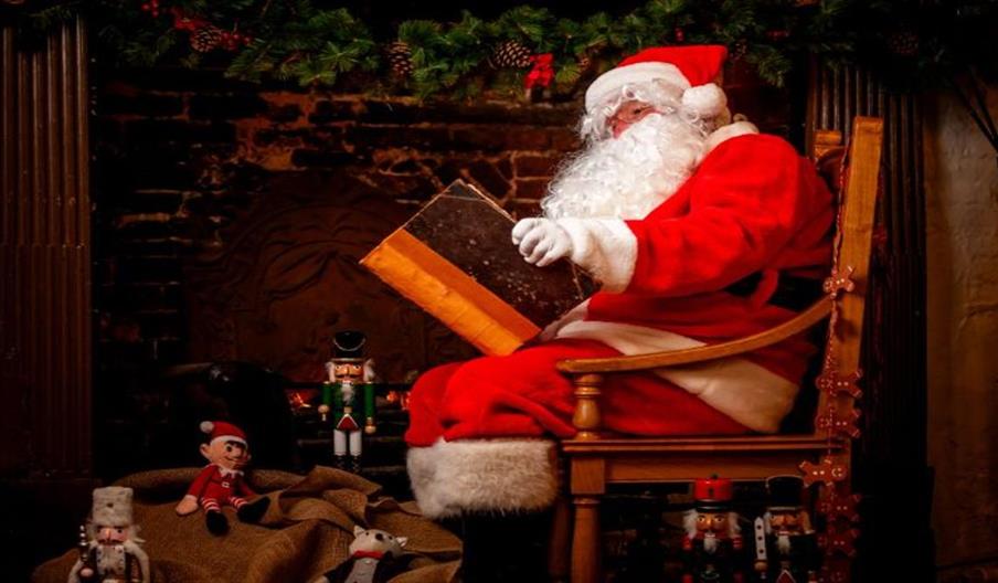 Santa Claus dressed in a traditional red suit with white trim sits in a wooden chair beside a fireplace, holding and reading a large book. The setting