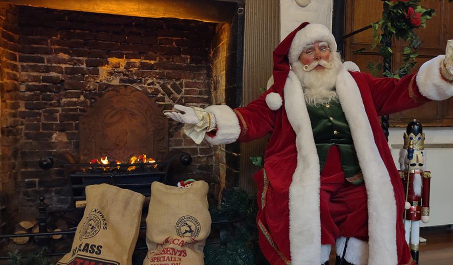 Santa sits, arms outstretched, beside a fireplace and some sacks of presents