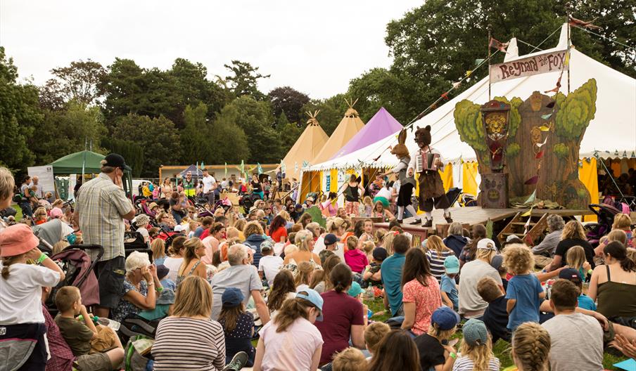 Image shows a crowd of people watching a performance featuring costumed characters, with a number of marquees behind the stage