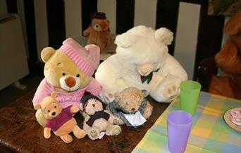 A selection of teddies of different sizes, on a wooden table, with a picnic off to the side