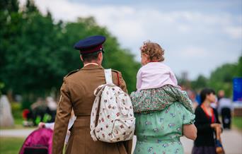 A soldier in uniform walks next to a woman in a vintage dress, with a young child on her shoulders