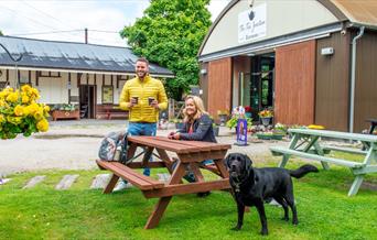 A male and female sitting at a wooden picnic bench with a dog, outside the Tea Junction Café and Manifold Information Point