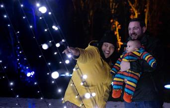 String lights are hanging down to the left. To the right, a woman wearing a yellow coat points at the lights while smiling. Next to her is a smiling m