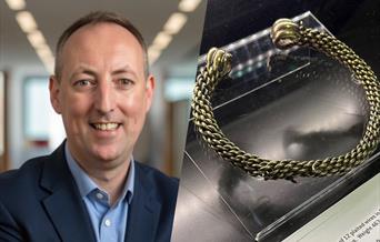 A picture of a man in a blue suit next to the Torc - a curved piece of metal
