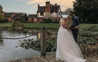 Image shows a bride and groom, at dusk, looking over the lake at the farm house on their big day