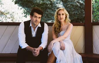 A man dressed in black and white and a woman in a long white dress, sitting in a wooden bandstand or similar
