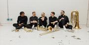 a group of 5 people casually seated on the floor, wearing black outfits, smiling and chatting with brass instruments laid out in front of them
