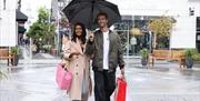 Two shoppers enjoy a day out at McArthurGlen West Midlands, carrying shopping bags and an umbrella as the rain falls