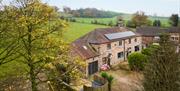 A cottage with solar panels, captured from the air, with green fields and tall trees behind it