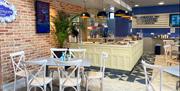 An empty cafe awaiting guests, with a host of tables and chairs and a bar with cakes. The image shows two of the walls, one of which is exposed brick,