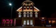 An image taken at night, across the road from an old red brick building with large windows.