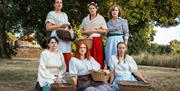 Six ladies outside on the grass, holding wicker baskets, looking quite serious