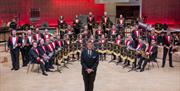 A band leader stands facing the camera, with a huge band behind him in rows, holding their instruments