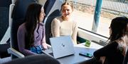 A group of young people on a train in a station, chatting. A laptop is open on the table, with a mobile phone next to it. It's a sunny day.
