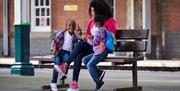 A family, with a mum and two kids, waits on the station platform for their train. They are all wearing colourful clothes and look like they are headin