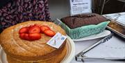 A  table display of homemade cakes at a community event, featuring a gluten‑free Victoria sponge topped with strawberries and a vegan chocolate loaf c
