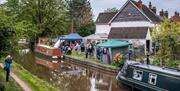 A  canalside garden event with people gathered under gazebos beside two moored narrowboats. One boat is painted red and green with the name "The Admir