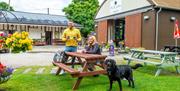 A male and female sitting at a wooden picnic bench with a dog, outside the Tea Junction Café and Manifold Information Point