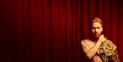 A topless man, with a Spanish matador's jacket draped over his shoulder, looks at the camera as he stands in front of a red curtain