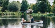 Image shows a couple taking a romantic boat trip, in their wedding outfits, across the lake. There's greenery all around the lake, and it's a cloudy d