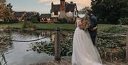 Image shows a bride and groom, at dusk, looking over the lake at the farm house on their big day