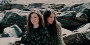 Two ladies, deep in thought, as they sit on some rocks on a beach